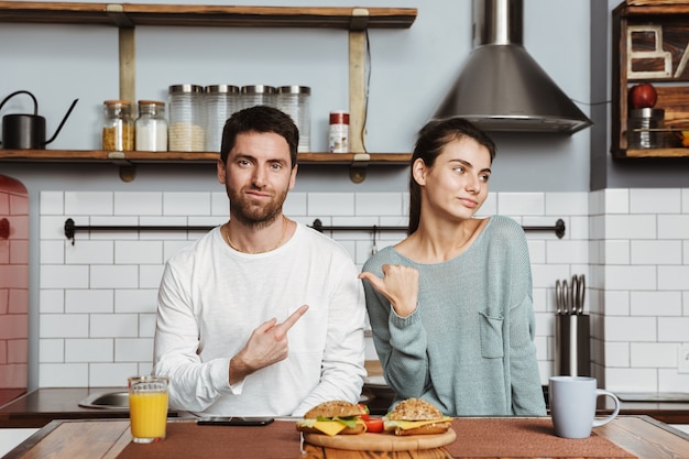 Jong koppel zittend aan de keuken tijdens het ontbijt thuis, wijzende vingers naar elkaar