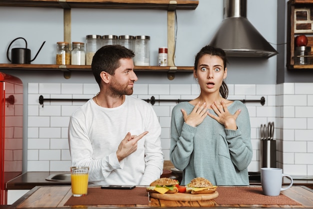 Jong koppel zittend aan de keuken tijdens het ontbijt thuis, wijzende vingers naar elkaar