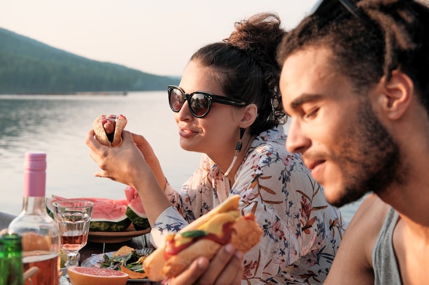 Foto jong koppel zittend aan tafel hotdogs eten op de natuur buitenshuis