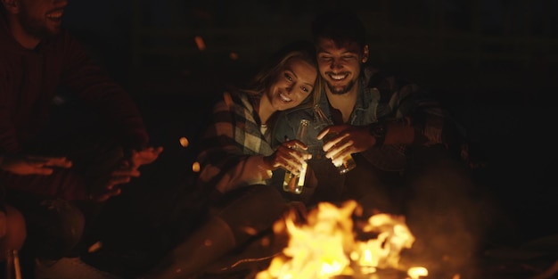 Jong koppel zittend met vrienden rond kampvuur op het strand 's nachts bier drinken