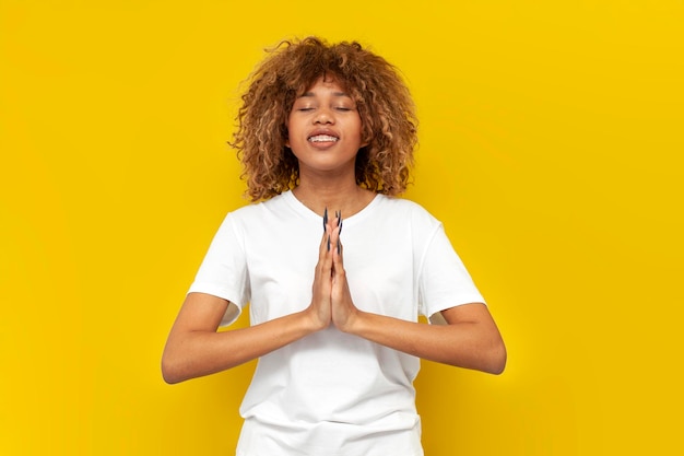jong krullend amerikaans meisje met beugels bidden en hopen op geluk op gele geïsoleerde achtergrond