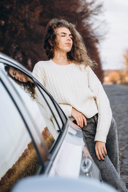 Foto jong krullend meisje in een witte trui tegen de achtergrond van de herfst natuur en haar eigen auto