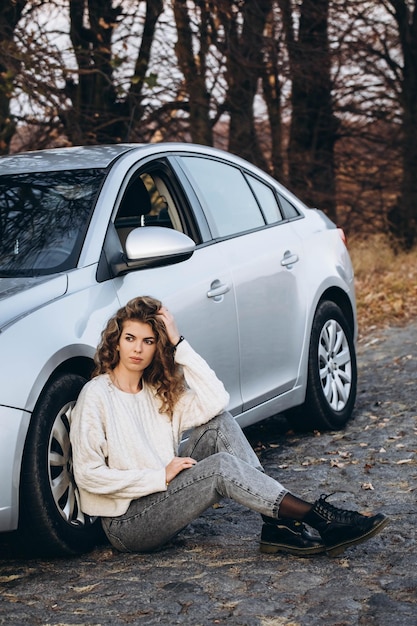 Foto jong krullend meisje in een witte trui tegen de achtergrond van de herfst natuur en haar eigen auto