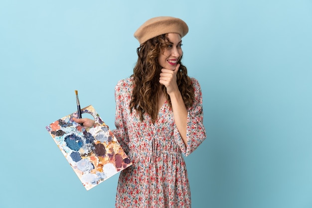 Jong kunstenaarsmeisje dat een palet houdt dat op blauwe muur wordt geïsoleerd die naar de kant kijkt en glimlacht