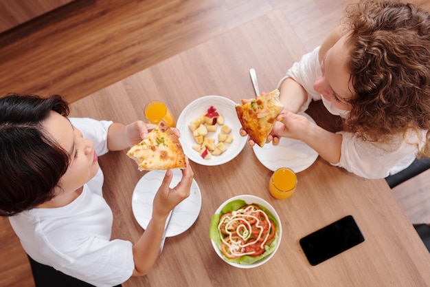 Jong lesbisch koppel dat pizzapunten eet met zelfgemaakte salade tijdens het eten thuis, uitzicht vanaf de top