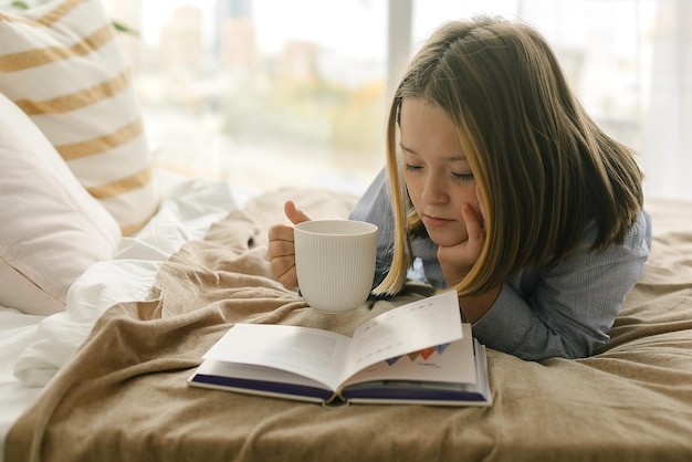 Jong meisje 8-9 jaar warme drank drinken, boek lezen, weekend doorbrengen in gezellig bed. Concept van herfst- of winterweekend