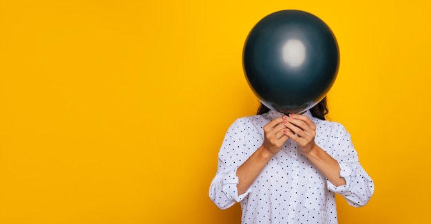 Jong meisje bedekte haar gezicht met behulp van zwarte ballon geïsoleerd op gele achtergrond