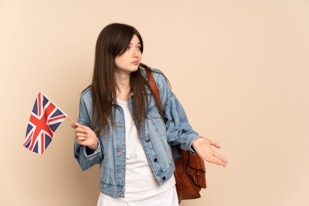 Jong meisje dat een vlag van het Verenigd Koninkrijk houdt die op beige wordt geïsoleerd met verrassingsuitdrukking terwijl zij kant kijkt
