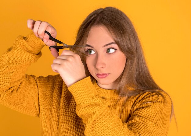 Jong meisje dat haar haar met schaar snijdt, die doen schrikken of doen schrikken, gele muur kijkt