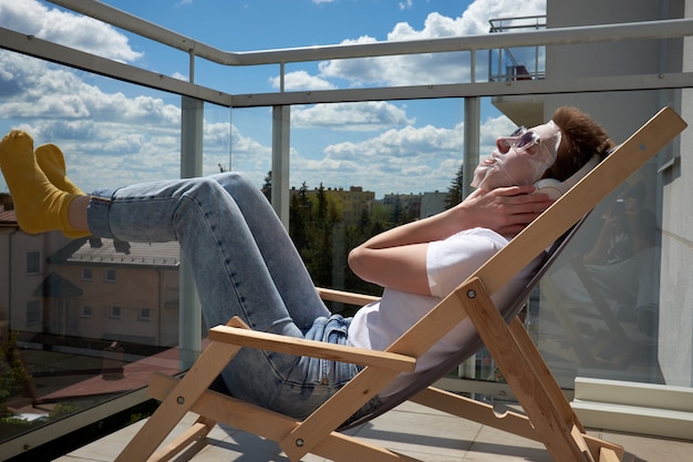 Jong meisje dat met leuk kapsel gezichtsmasker op gezicht toepast, muziek op hoofdtelefoons luistert en op het balkon zit.