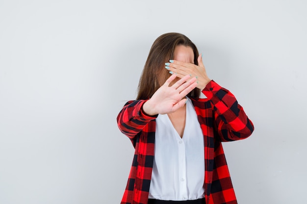 Jong meisje dat ogen bedekt met palm, stopgebaar toont in geruit hemd, blouse en boos kijkt, vooraanzicht.