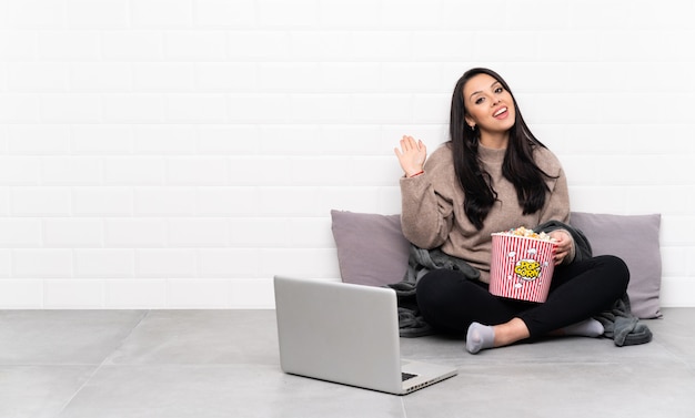 Jong meisje die een kom popcorns houden en een film in laptop tonen die met hand met gelukkige uitdrukking groeten