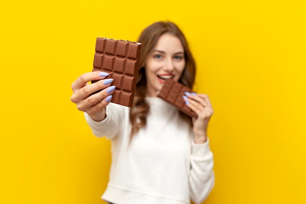 jong meisje eet en bijt grote chocoladereep en biedt stuk aan u op gele geïsoleerde achtergrond