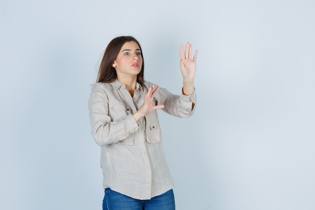 Jong meisje in beige shirt, jeans die handen opheft om te stoppen en er bang uit te zien, vooraanzicht.