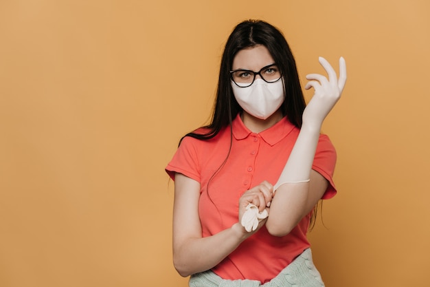 Jong meisje in chirurgische masker en bril, gekleed in een roze t-shirt, het aantrekken van beschermende handschoenen, ziet er zelfverzekerd uit, bescherming tegen pandemie van het coronavirus, quarantaine thuis. Covid-19 concept.