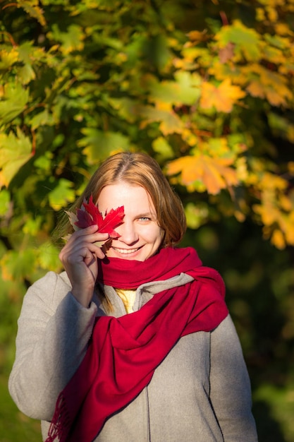 Jong meisje in een grijze jas en een rode sjaal met een esdoornblad in haar hand op een herfstachtergrond