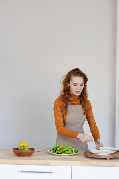 Jong meisje in een schort maakt een gezonde salade van groenten en kruiden in haar keuken