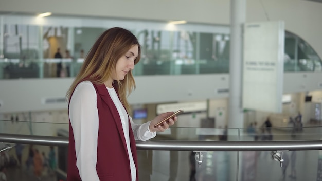 Foto jong meisje in elegante kleding staat en typt in de smartphone op de luchthaven.