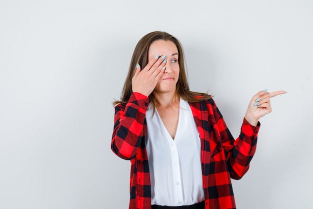 Jong meisje in geruit hemd, blouse die de ogen bedekt met de hand, naar de zijkant wijst en er attent uitziet, vooraanzicht.