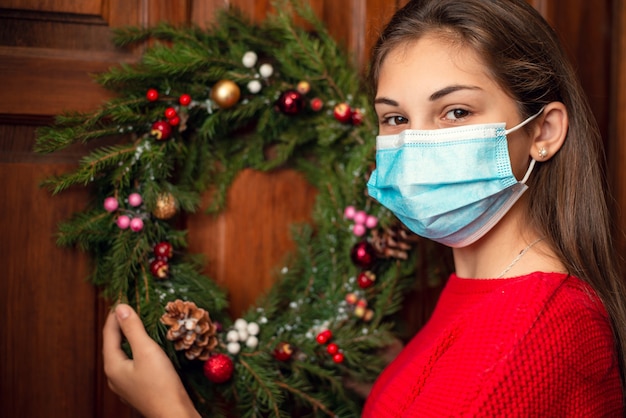 Jong meisje in medisch masker dat zich dichtbij de deur van haar huis bevindt en een zelfgemaakte Kerstmiskroon hangt