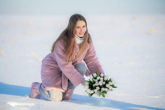 Jong meisje in roze jas die een boeket witte tulpen van de sneeuw op een winterdag ophaalt