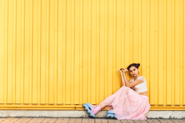 Jong meisje in roze rok en rolschaatsen zit op stoep tegen gele muur achtergrond en handen maakt hart