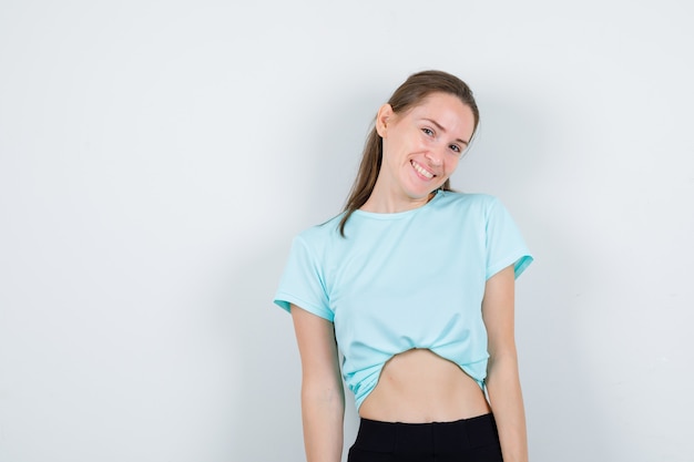 Jong meisje kijkt naar de camera in turquoise t-shirt, broek en ziet er vrolijk uit, vooraanzicht.