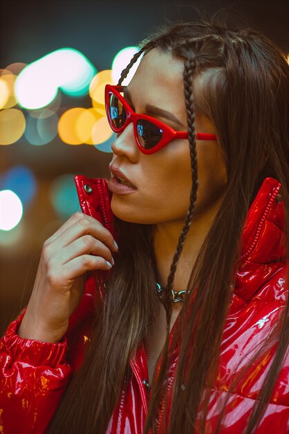 Jong meisje met bril buiten mode portret met gevlochten haar draagt rode donsjack en mode op neon straatverlichting