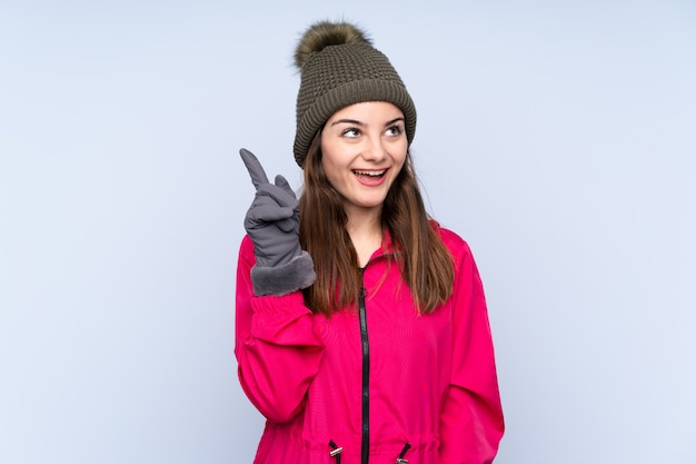 Jong meisje met de winterhoed dat op blauw wordt geïsoleerd dat de oplossing wil realiseren terwijl het opheffen van een vinger