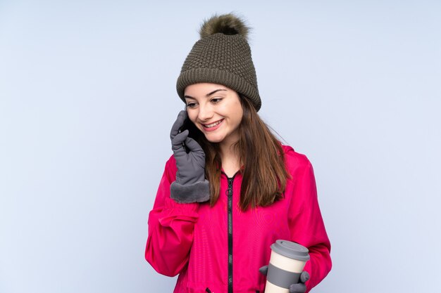 Jong meisje met de winterhoed dat op blauwe holdingskoffie wordt geïsoleerd om weg te halen en mobiel