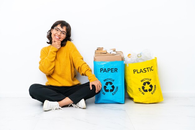 Jong meisje met een zak vol plastic en papier zittend op de vloer geïsoleerd op een witte achtergrond met een bril en glimlachend