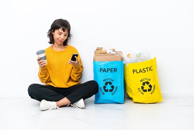 Jong meisje met een zak vol plastic en papier zittend op de vloer geïsoleerd op een witte achtergrond met koffie om mee te nemen en een mobiel