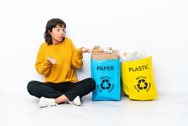 Jong meisje met een zak vol plastic en papier zittend op de vloer geïsoleerd op een witte achtergrond met verrassende gezichtsuitdrukking