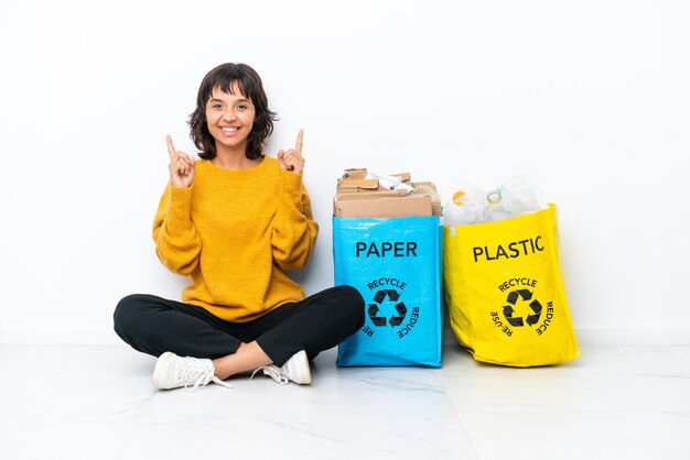 Jong meisje met een zak vol plastic en papier zittend op de vloer geïsoleerd op een witte achtergrond wijzend op een geweldig idee