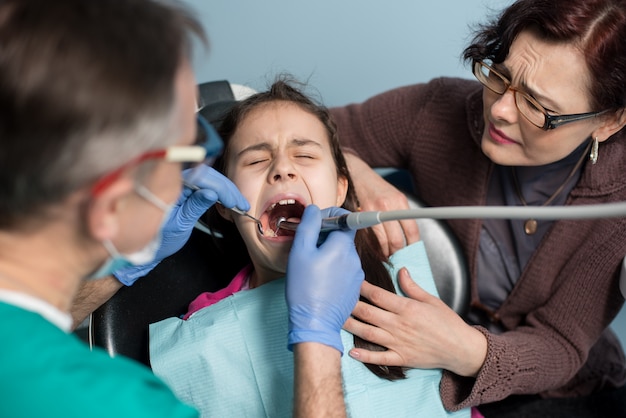 Jong meisje met haar moeder op het eerste tandbezoek. Hogere pediatrische tandarts die geduldige meisjestanden behandelen