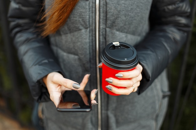 Jong meisje met koffie in rode kop en zwarte smartphone in hand buitenshuis