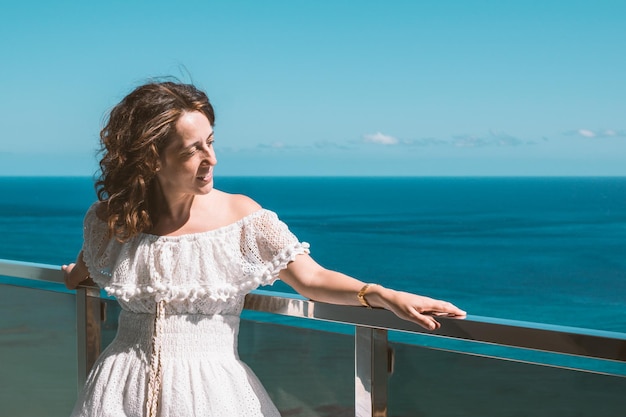 Jong meisje met lang bruin haar glimlachend in een witte zomerjurk op een hotelbalkon kijkend naar de kant met de blauwe zee op de achtergrond