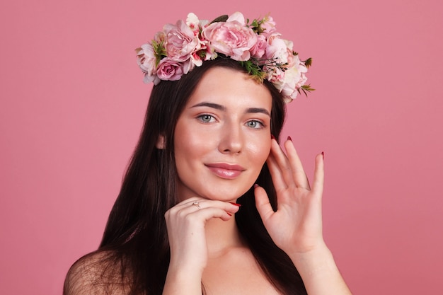 Jong meisje met natuurlijk haar en schone huid in een krans van bloemen op een roze achtergrond