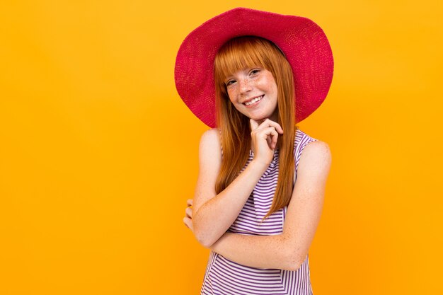 Jong meisje met rood haar en roze hoed glimlacht en hoopt op iets geïsoleerd op geel