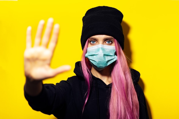 Jong meisje met roze haar en blauwe ogen, die medisch griepmasker, trui met capuchon en beanie-hoed draagt, stopgebaar op gele muur toont.