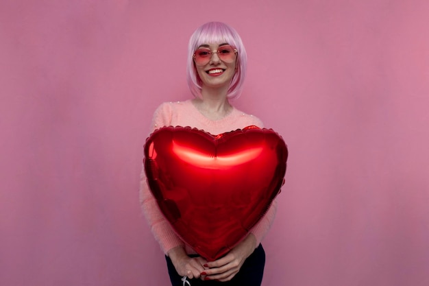 Jong meisje met roze haar houdt een hartballon vast en glimlacht op roze achtergrond vrouw op Valentijnsdag