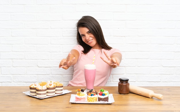 Jong meisje met veel verschillende minicakes die en overwinningsteken glimlachen tonen