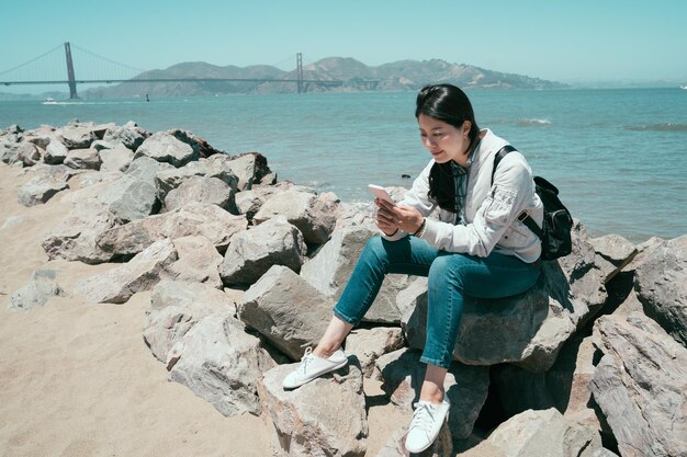 jong meisje reiziger zittend op steen in de buurt van de oceaan met behulp van mobiele telefoon chatten SMS-bericht op zonnige dag buiten ontspannen genieten van zonlicht. rode golden gate bridge op de achtergrond met blauwe lucht.