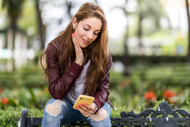 Jong meisje schrijft een bericht met haar smartphone zittend op een bankje in het parkformaat met kopieerruimte close-up