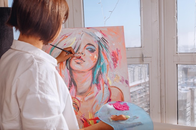 Jong meisje staat met haar rug en richt zich op het tekenen van een helder portret van een meisje in niet-standaard kleuren in een kunststudio in de buurt van een raam met uitzicht op de stad
