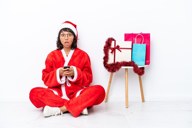 Jong meisje viert Kerstmis zittend op de vloer geïsoleerd op een witte achtergrond verrast en een bericht verzenden