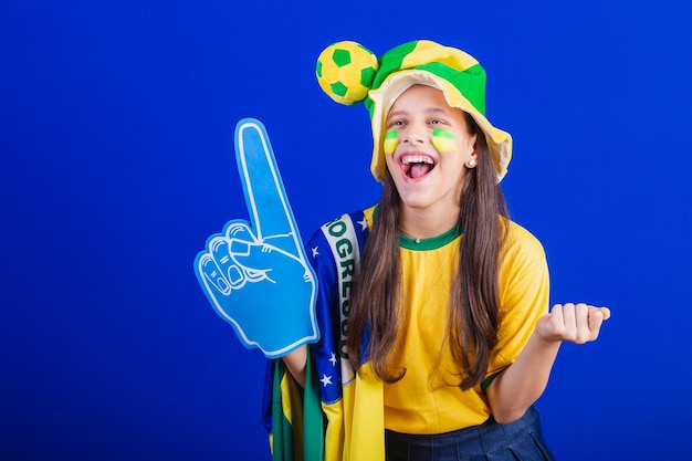 Jong meisje voetbalfan uit Brazilië gekleed in hoed en vlag met behulp van foam finger feesten geweldig