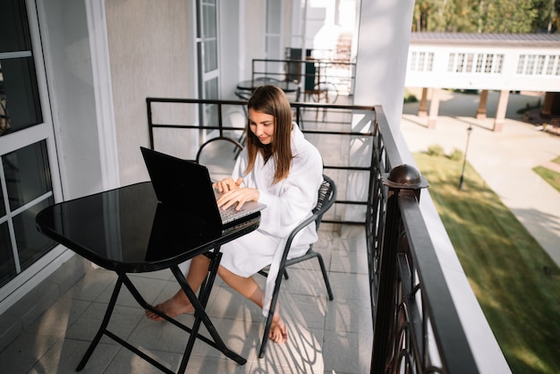 Jong meisje werkt op haar laptop op het balkon in een wit gewaad