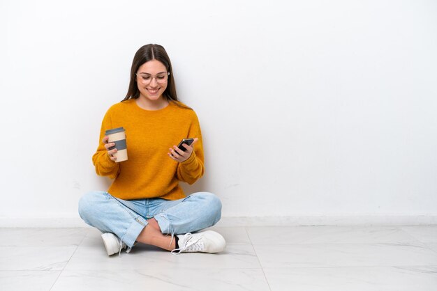 Jong meisje zittend op de vloer geïsoleerd op een witte achtergrond met koffie om mee te nemen en een mobiel