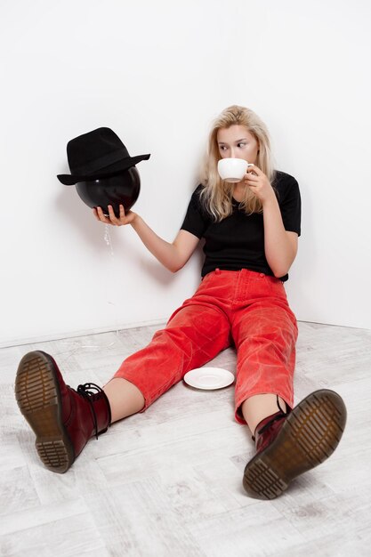 Jong mooi blond meisje met zwarte ballon in hoed zittend op de vloer koffie drinken over witte muur